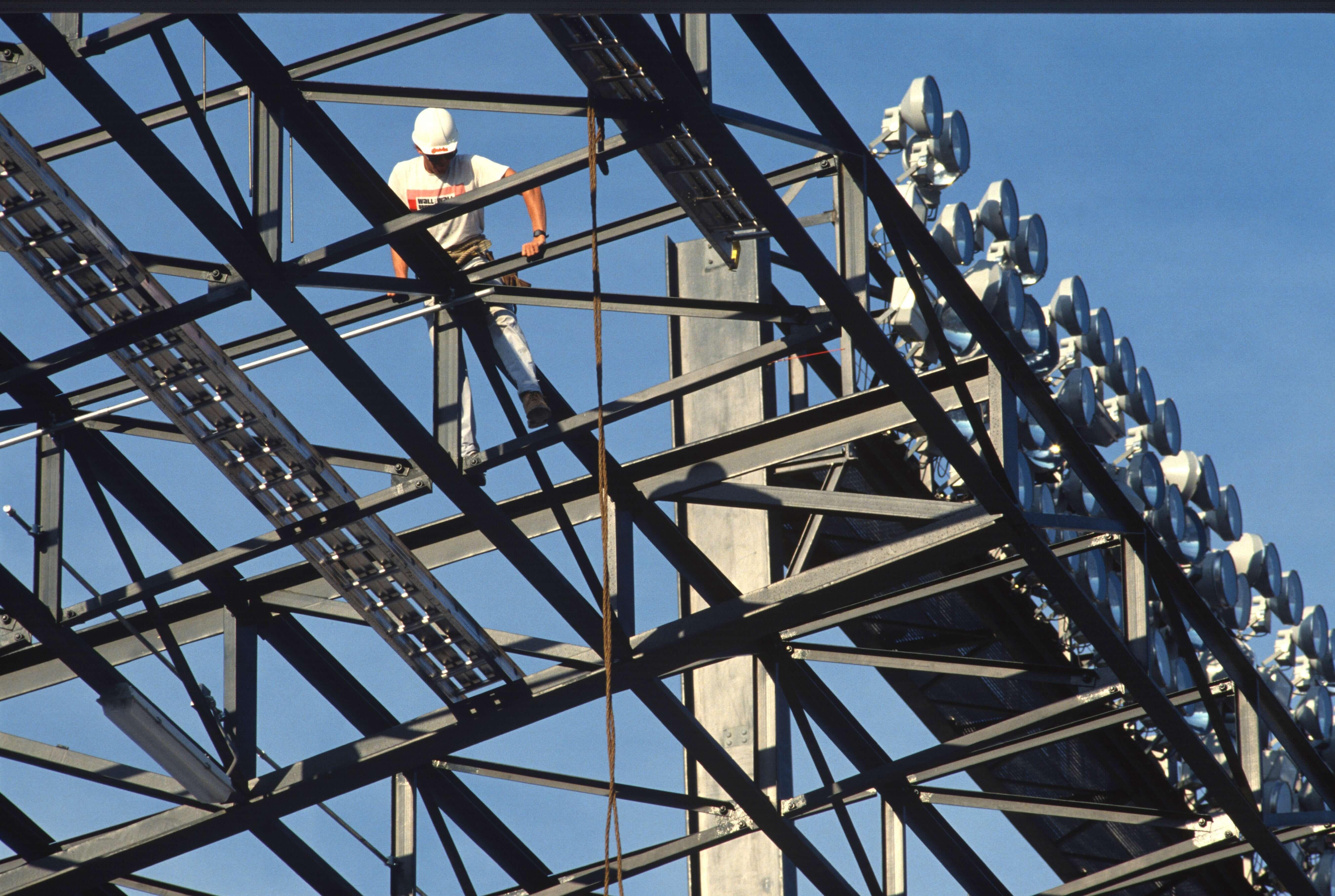Construction worker walking across steel beams while installing stadium light structures against a blue sky.