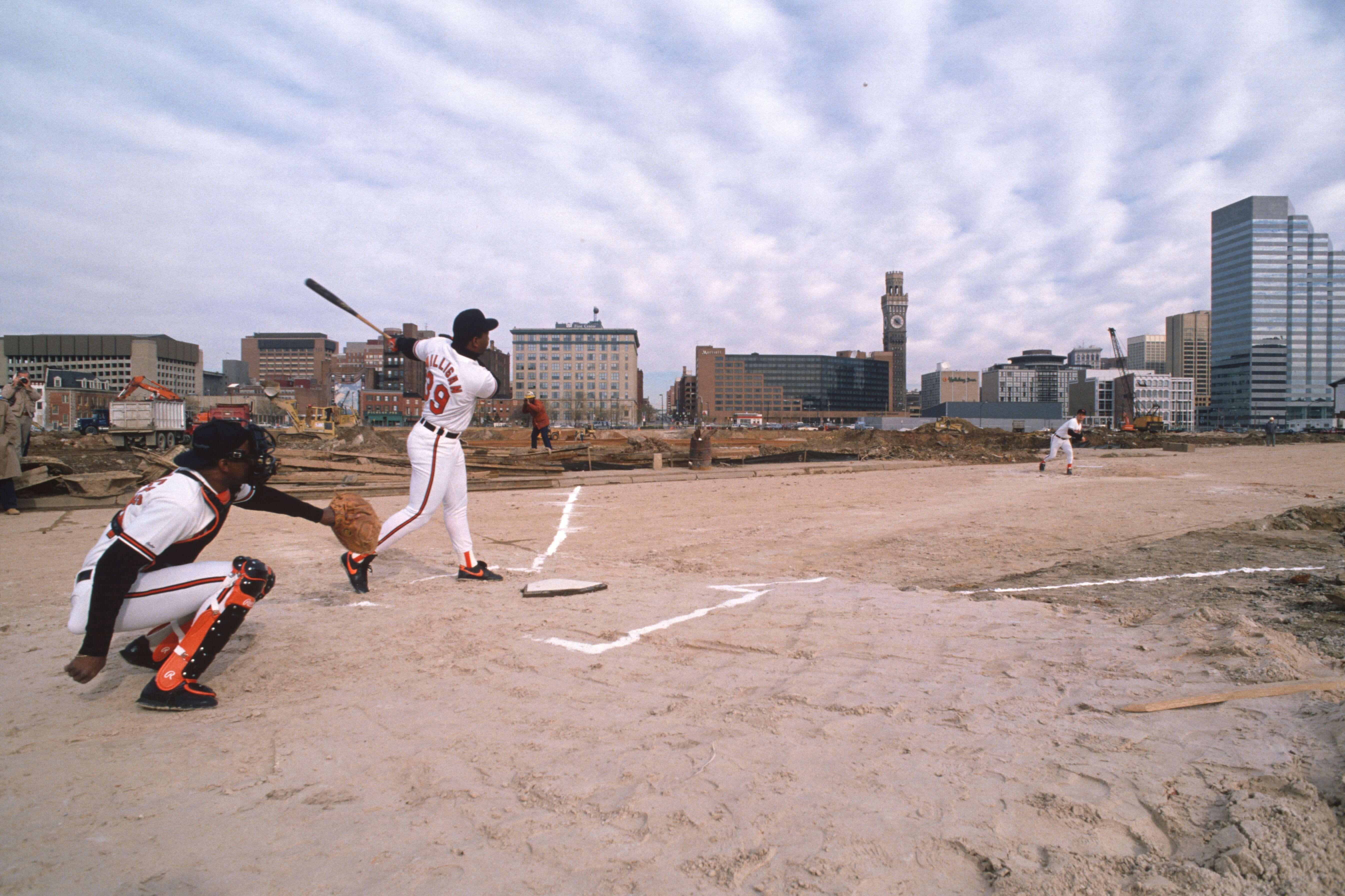 Baltimore Orioles players batting and fielding on a dirt field at the Camden Yards construction site with the city skyline behind them.