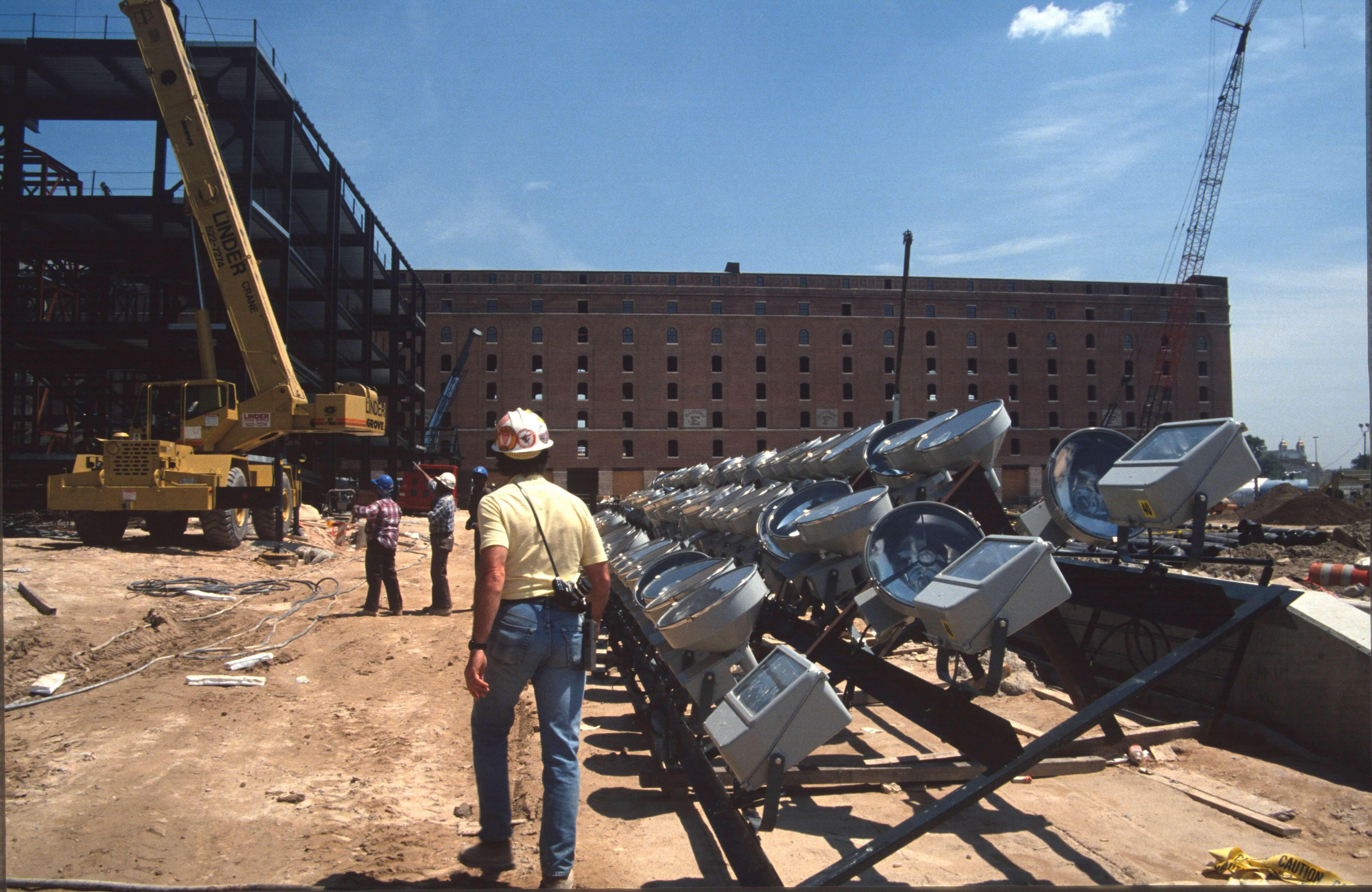 Stadium light fixtures lined up on the ground before installation, with workers and cranes in the background.