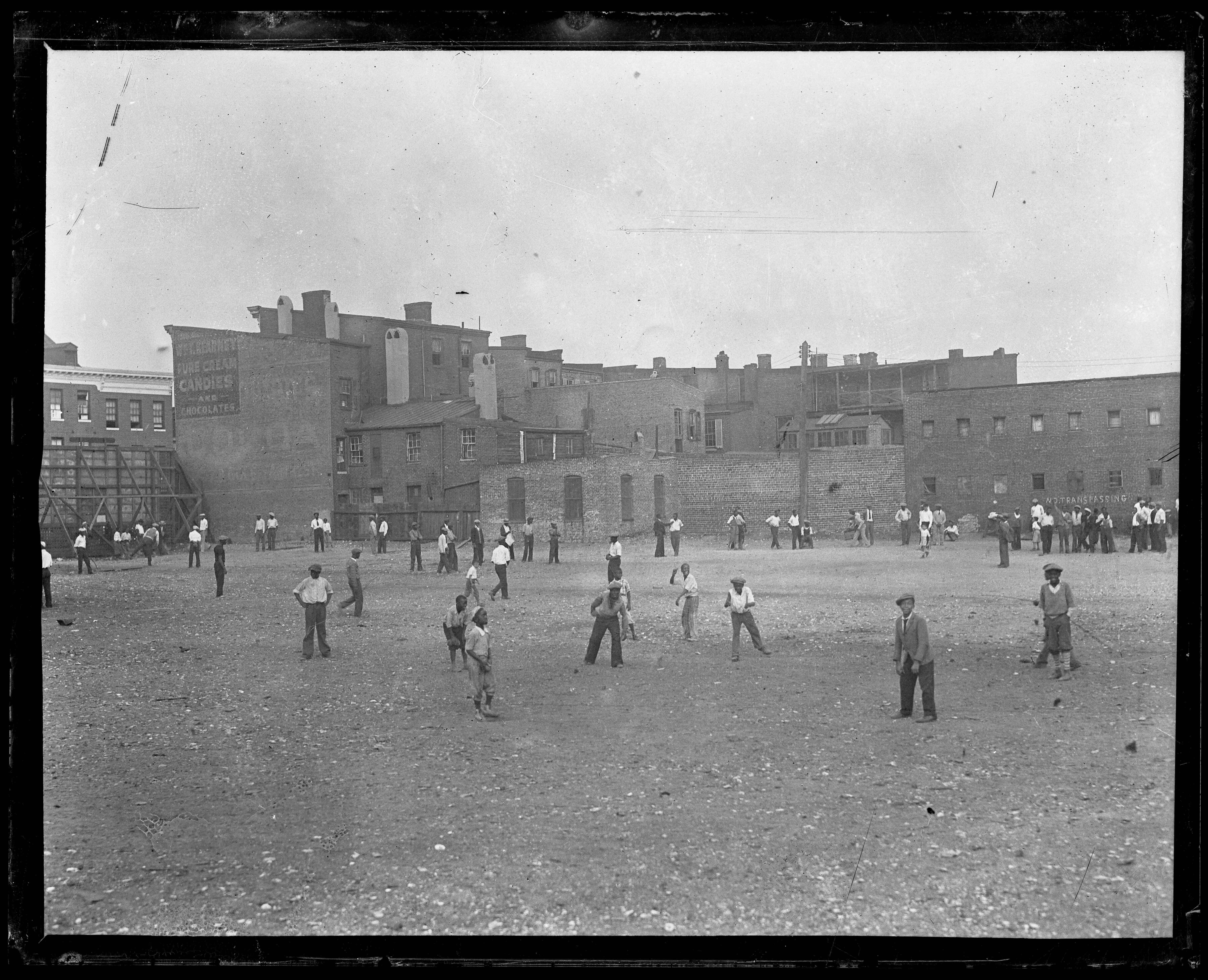 Large group of men and boys playing and watching a neighborhood baseball game in an open dirt lot surrounded by brick buildings.