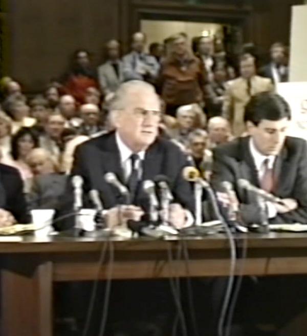 Man testifies at a government hearing table with multiple microphones as an audience looks on.