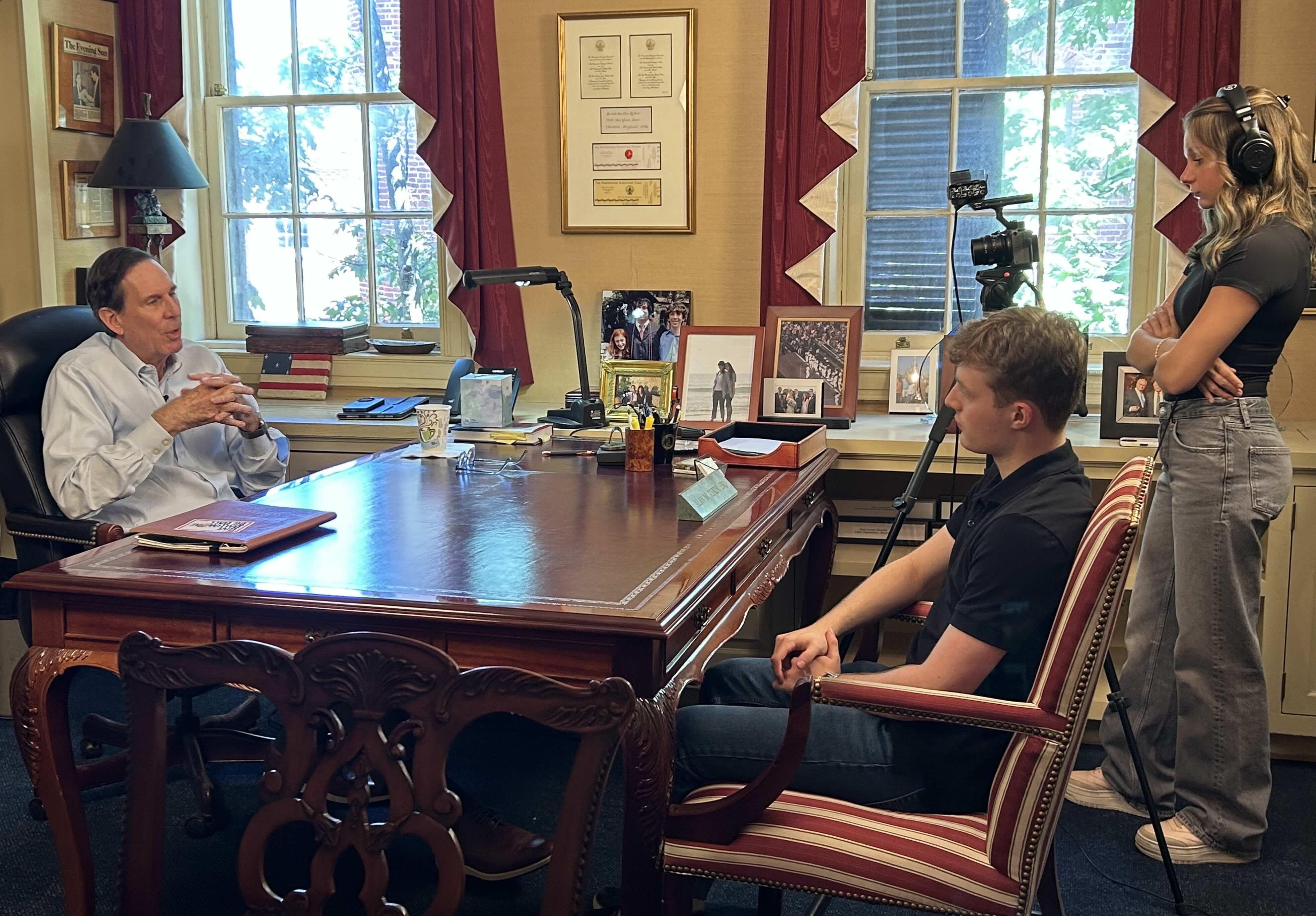 Man being interviewed at a desk while two people film with a camera and audio equipment.