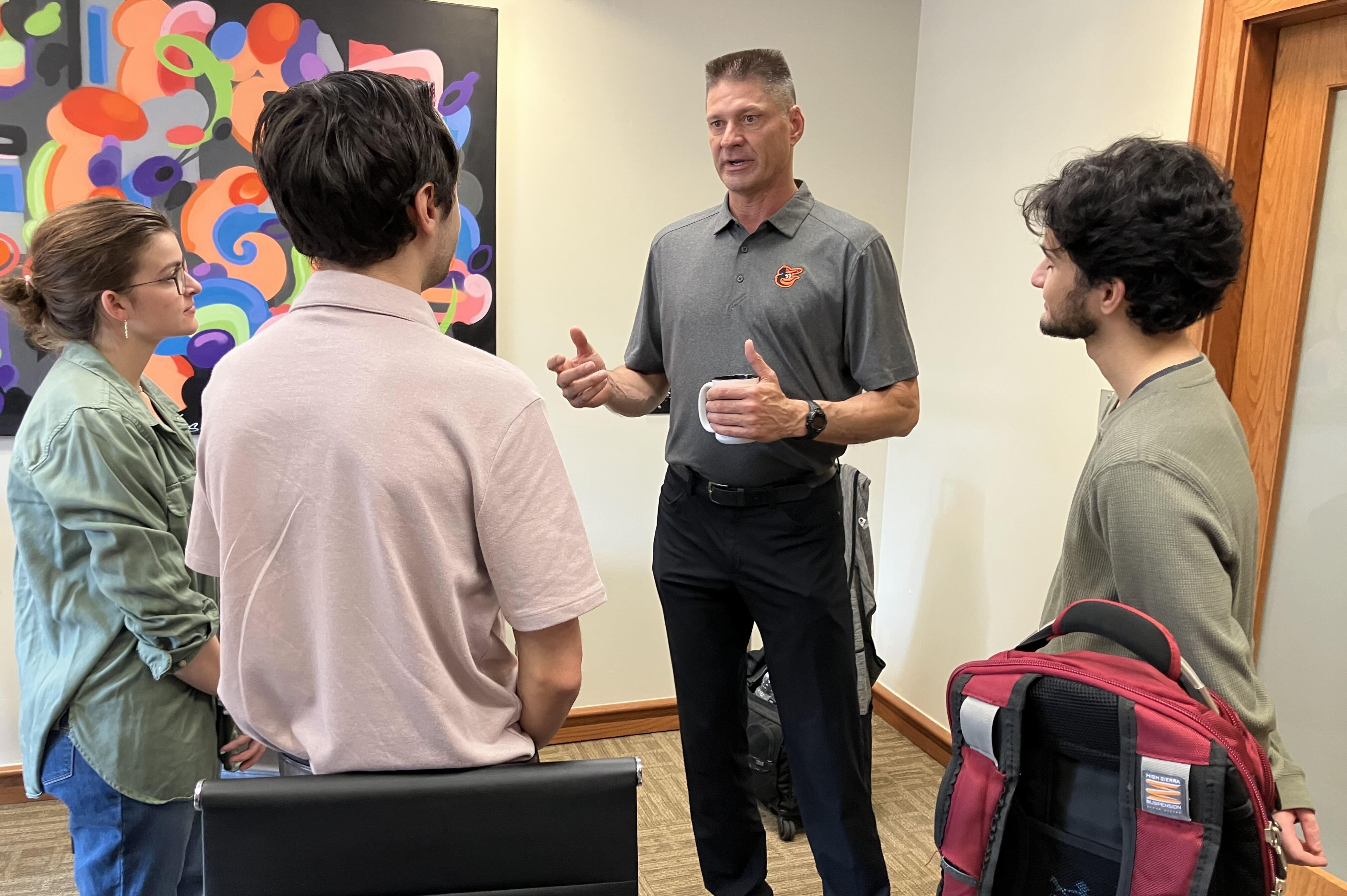 Man speaking with three young people standing in a small office during an informal discussion.