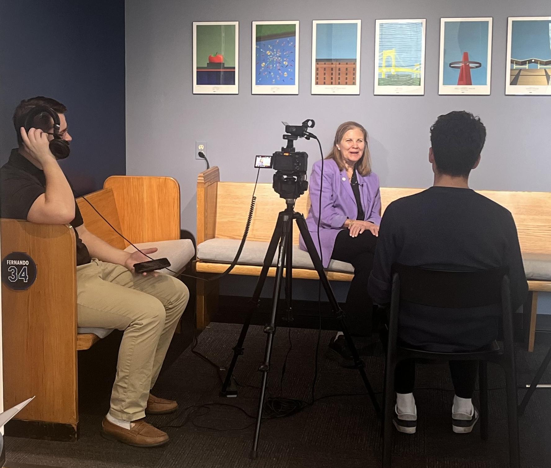 Woman seated on a bench being interviewed while a small crew records video and audio.