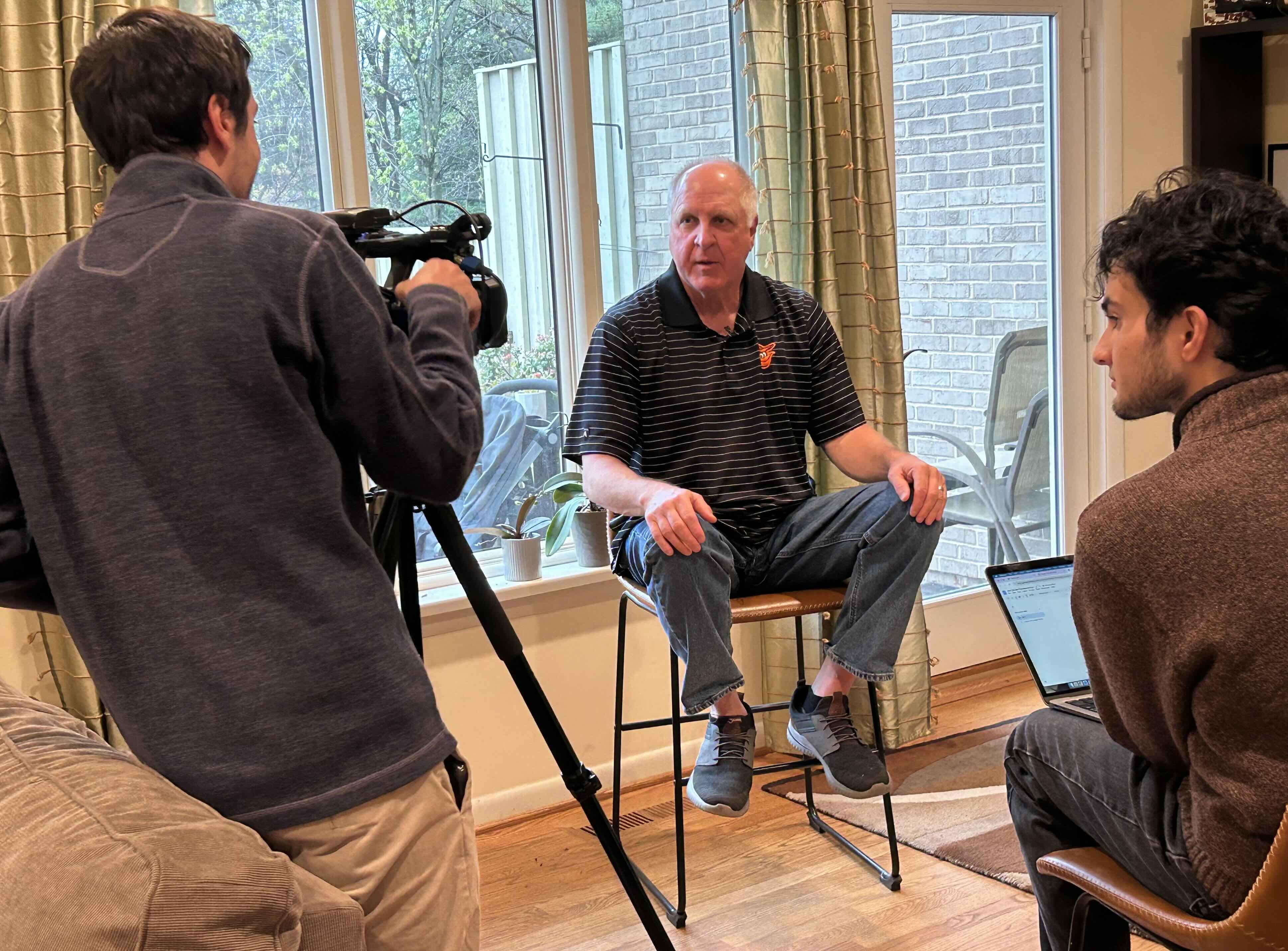 Man being interviewed in a living room while two people operate a camera and take notes.