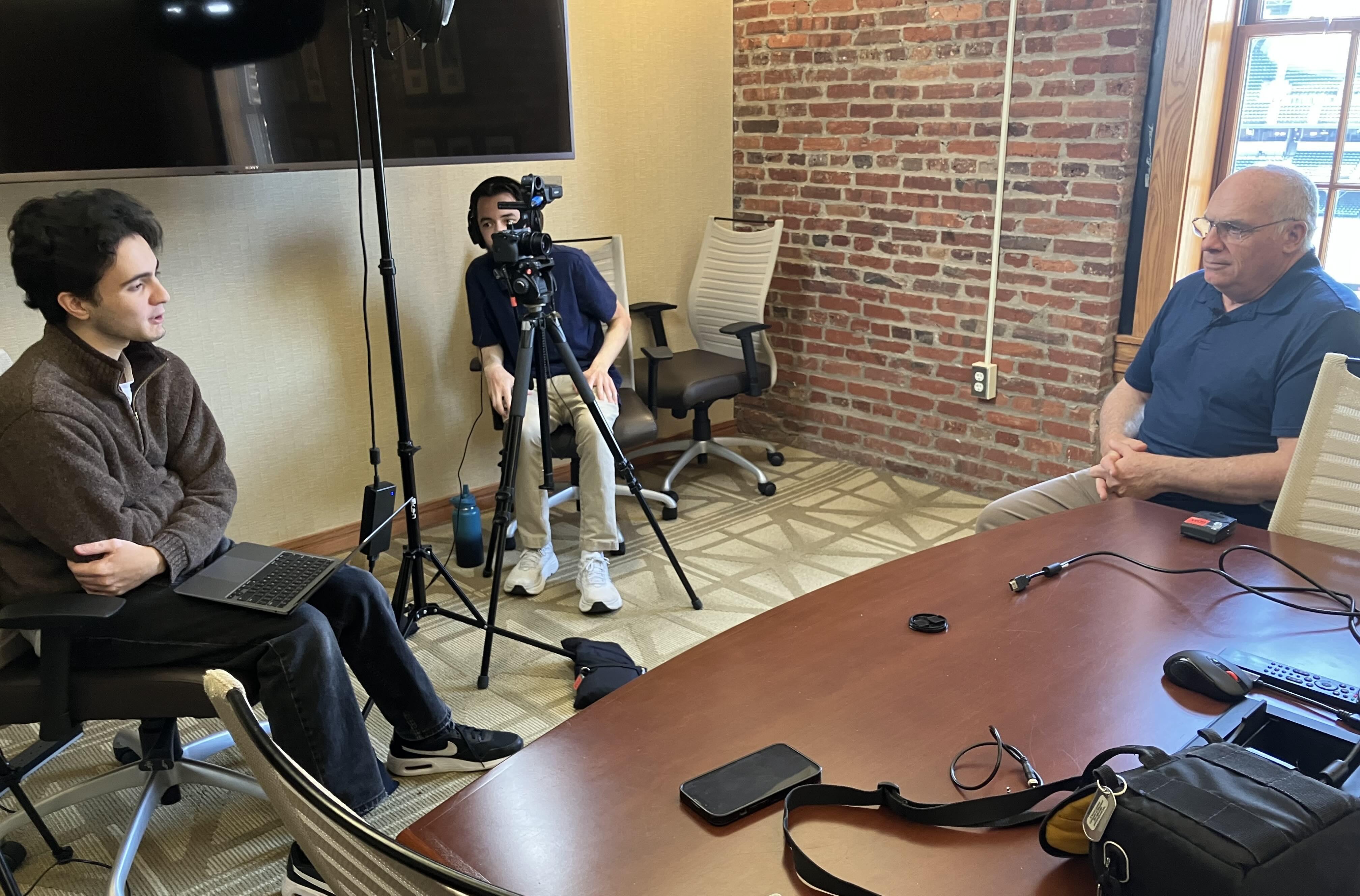 Man seated at a conference table being interviewed by two people with a camera.