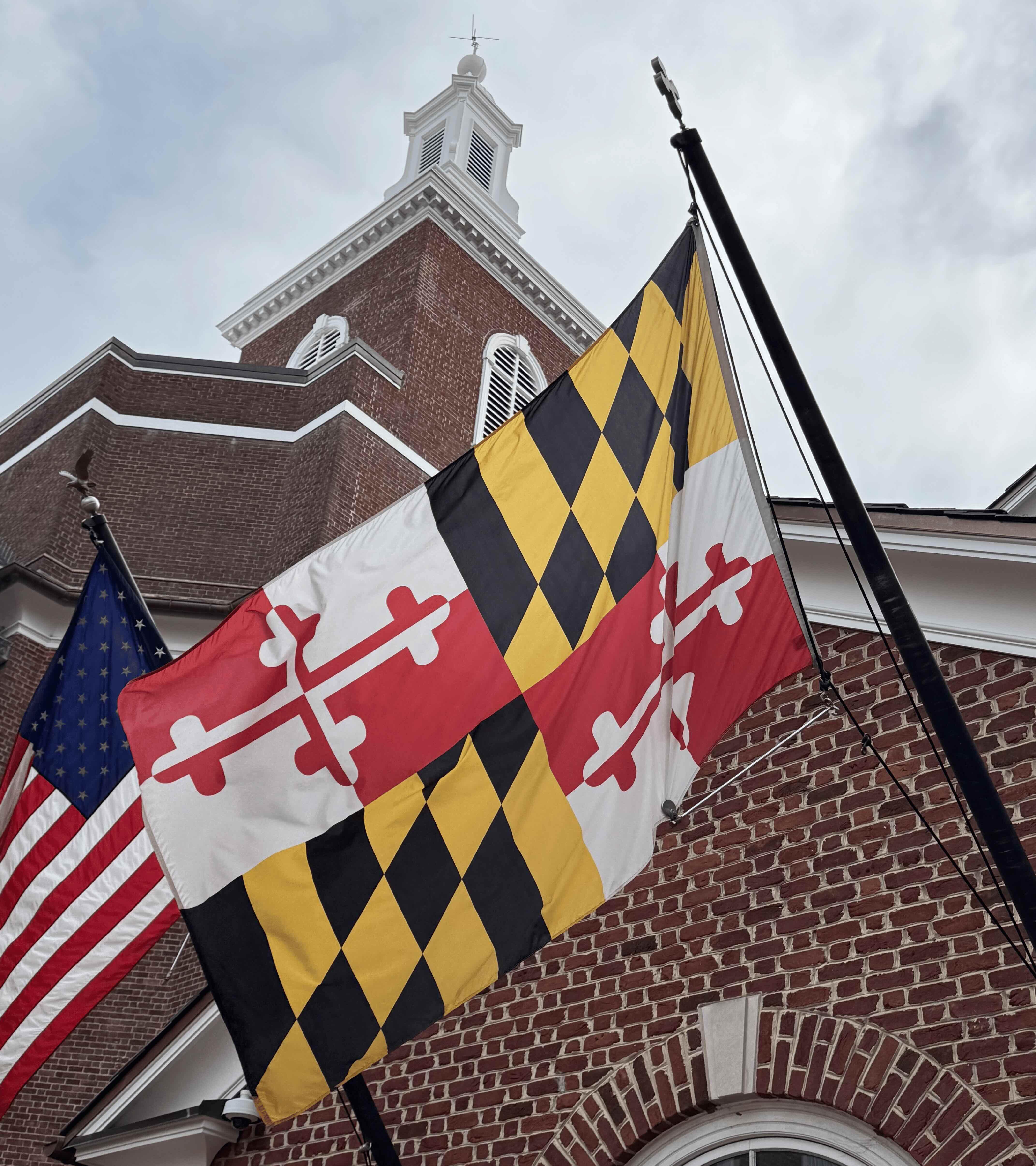 Yellow, red, and black Maryland flag waving in front of a brick building and the American flag